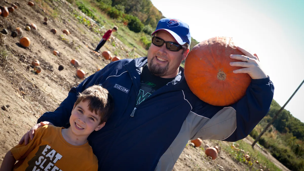 Dad carrying a pumpkin on his shoulder with his son in the pumpkin field